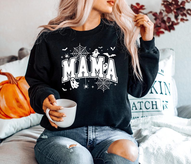 Op de afbeelding: Zwart sweatshirt met het woord "MAMA" in witte letters, versierd met spinnenwebben, vleermuizen, spinnen en geesten. Een ontwerp met Halloween-thema.