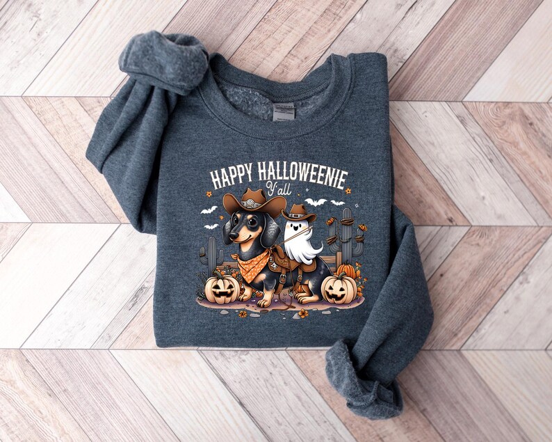 May include: A dark grey sweatshirt featuring the phrase "HAPPY HALLOWEENIE y'all" and a graphic of a dachshund in a cowboy hat and a ghost. The dachshund is wearing a cowboy hat and a bandana. Pumpkins and cacti are also in the graphic.