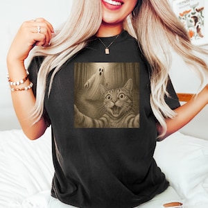 Może przedstawiać: Czarny t-shirt z sepiową grafiką kota robiącego selfie z duchem w lesie. Kot ma szeroko otwarte oczy i otwarte usta, wyglądając na zaskoczonego. Duch jest biały i przezroczysty. Koszulka jest w stylu casual.