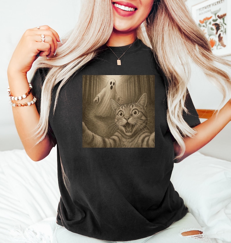 Może przedstawiać: Czarny t-shirt z sepiową grafiką kota robiącego selfie z duchem w lesie. Kot ma szeroko otwarte oczy i otwarte usta, wyglądając na zaskoczonego. Duch jest biały i przezroczysty. Koszulka jest w stylu casual.