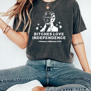 May include: Dark gray t-shirt with a white graphic of Thomas Jefferson and the text "BITCHES LOVE INDEPENDENCE - Thomas Jefferson 1776". The shirt is a crew neck and has short sleeves.