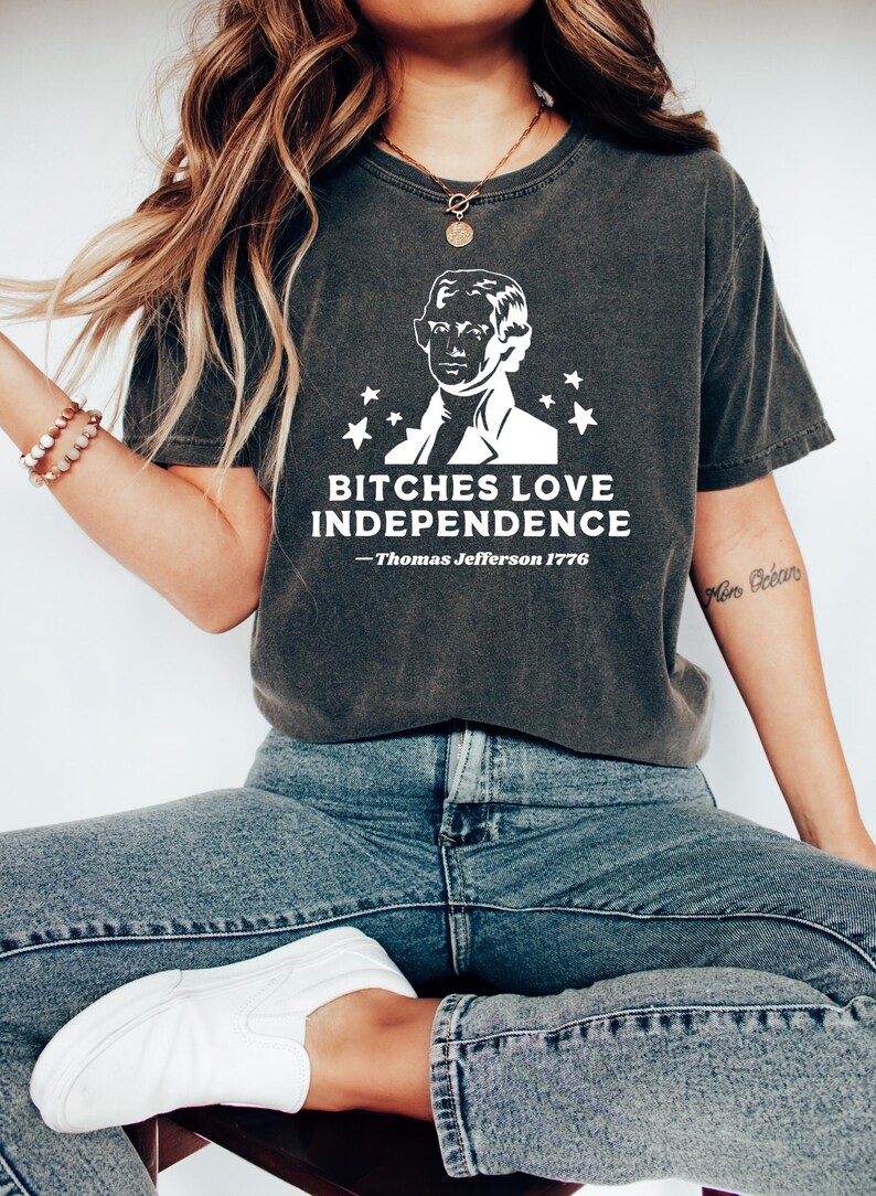 May include: Dark gray t-shirt with a white graphic of Thomas Jefferson and the text "BITCHES LOVE INDEPENDENCE - Thomas Jefferson 1776". The shirt is a crew neck and has short sleeves.
