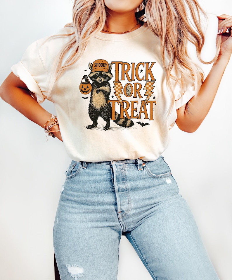 Peut inclure: T-shirt cr&egrave;me avec un motif d'Halloween. Le design comprend un raton laveur portant un chapeau et tenant une lanterne citrouille, avec les mots "Spooky Trick or Treat" en orange et marron.