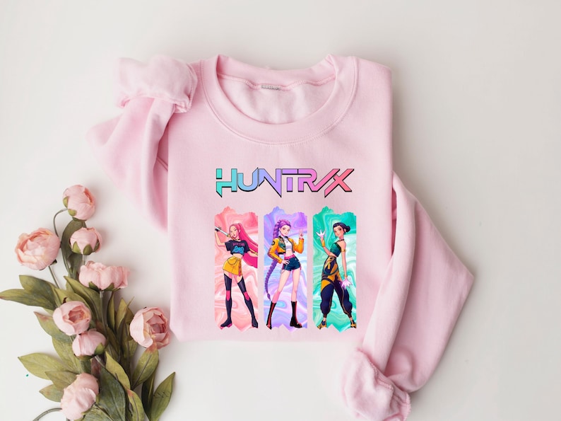 May include: A light pink sweatshirt featuring the word "HUNTRAX" in a colorful font. Below the text are three anime-style characters in colorful outfits. The sweatshirt is made of a soft material and is suitable for casual wear.