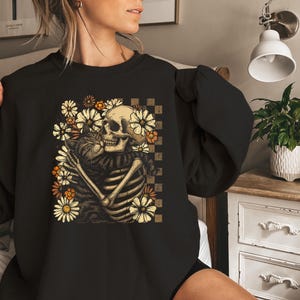 May include: Black sweatshirt featuring a skeleton embracing a cat, surrounded by daisies. The design includes a checkered background and is in shades of beige, orange, and brown. The sweatshirt is a crew neck style.