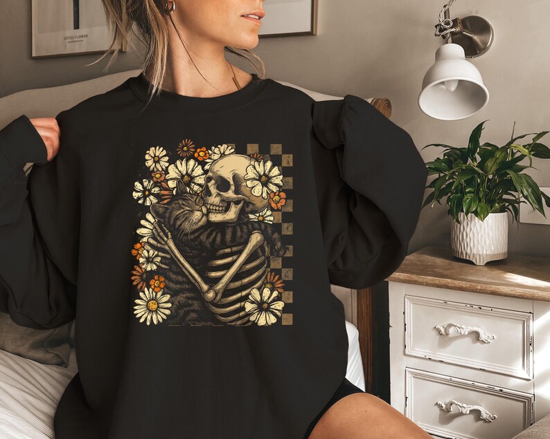 May include: Black sweatshirt featuring a skeleton embracing a cat, surrounded by daisies. The design includes a checkered background and is in shades of beige, orange, and brown. The sweatshirt is a crew neck style.