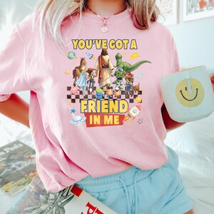 May include: A light pink t-shirt featuring a graphic print of a central figure walking with various animated characters, including a dinosaur and a cowboy. The text reads YOU'VE GOT A FRIEND IN ME, surrounded by space-themed icons like planets and rockets.