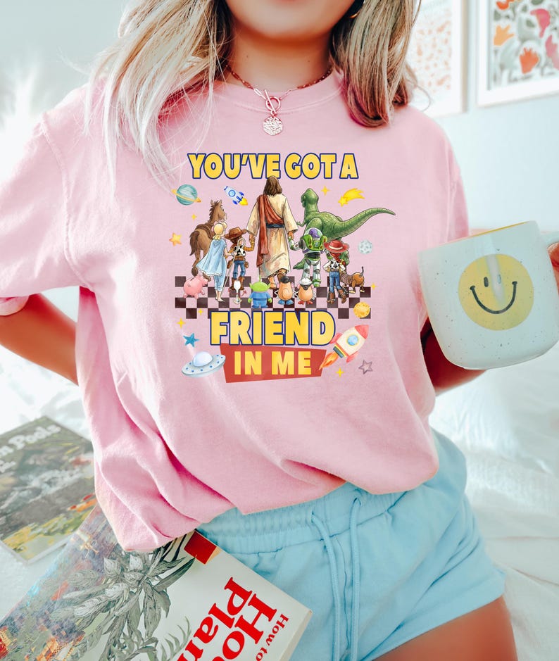 May include: A light pink t-shirt featuring a graphic print of a central figure walking with various animated characters, including a dinosaur and a cowboy. The text reads YOU'VE GOT A FRIEND IN ME, surrounded by space-themed icons like planets and rockets.