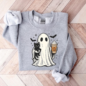 May include: A light grey sweatshirt with a cartoon ghost holding a black cat and a pumpkin spice latte. The ghost has two black eyes and is surrounded by bats and stars. The sweatshirt is made of a soft material.