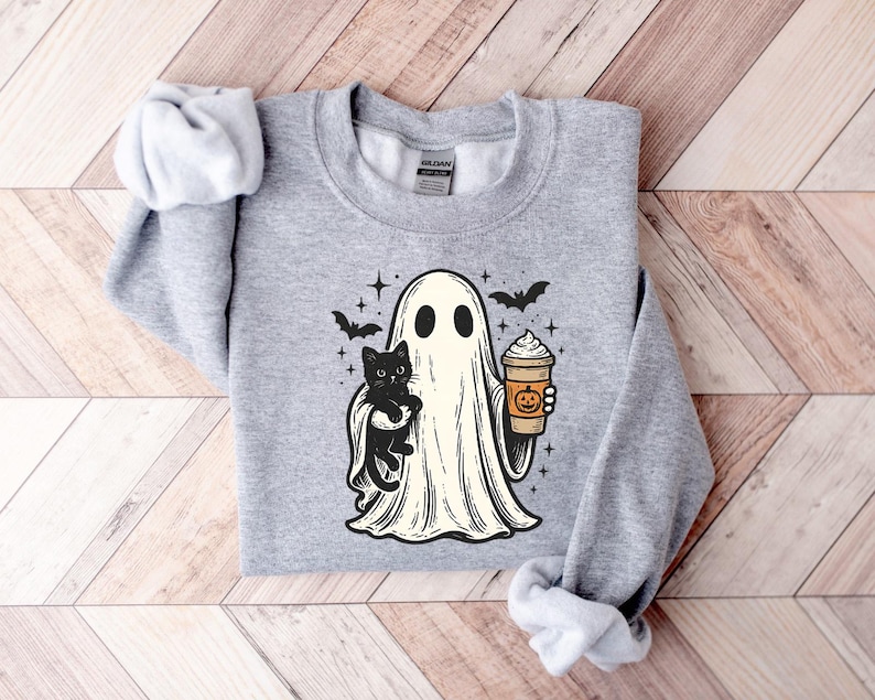 May include: A light grey sweatshirt with a cartoon ghost holding a black cat and a pumpkin spice latte. The ghost has two black eyes and is surrounded by bats and stars. The sweatshirt is made of a soft material.