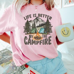 May include: Pink t-shirt with a graphic of a mountain scene with two tents, a campfire, and trees. The text "Life is better by the campfire" is printed on the shirt.