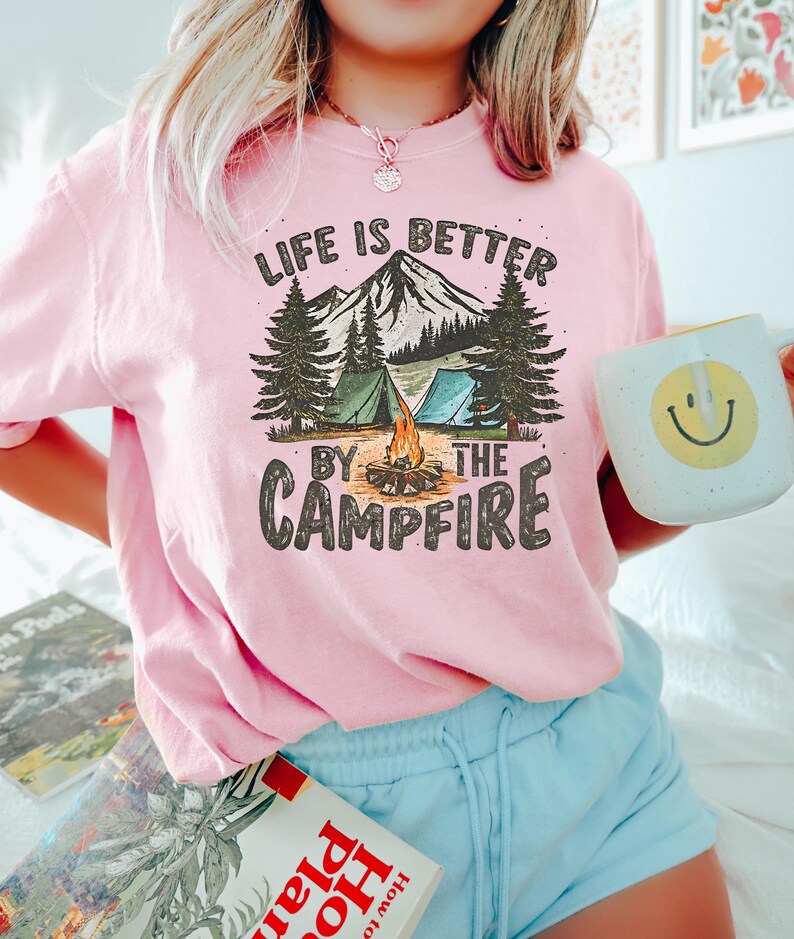May include: Pink t-shirt with a graphic of a mountain scene with two tents, a campfire, and trees. The text "Life is better by the campfire" is printed on the shirt.