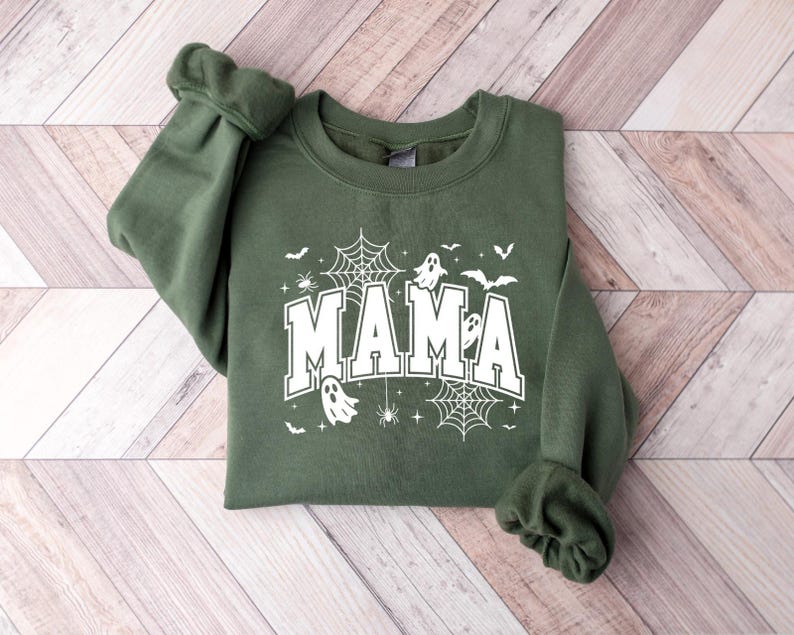Op de afbeelding: Een olijfgroene sweatshirt met het woord "MAMA" in witte blokletters. Het ontwerp bevat spinnenwebben, vleermuizen, spinnen en geesten, wat een Halloween-thema cre&euml;ert. De mouwen zijn opgerold.