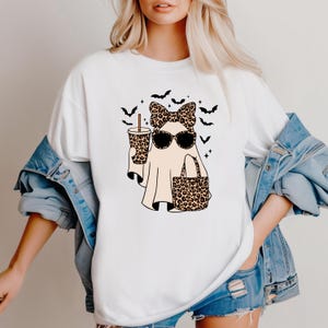 May include: White t-shirt featuring a cartoon ghost with sunglasses, a bow, and a tote bag, all in a leopard print design. The ghost is holding a coffee cup with the same print. Black bats and stars are also featured in the design.