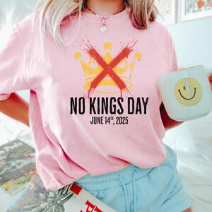May include: Pink t-shirt with the text "NO KINGS DAY" and the date "JUNE 14TH, 2025". The design includes a yellow crown crossed out with a red "X". A white mug with a yellow smiley face is held in the hand.