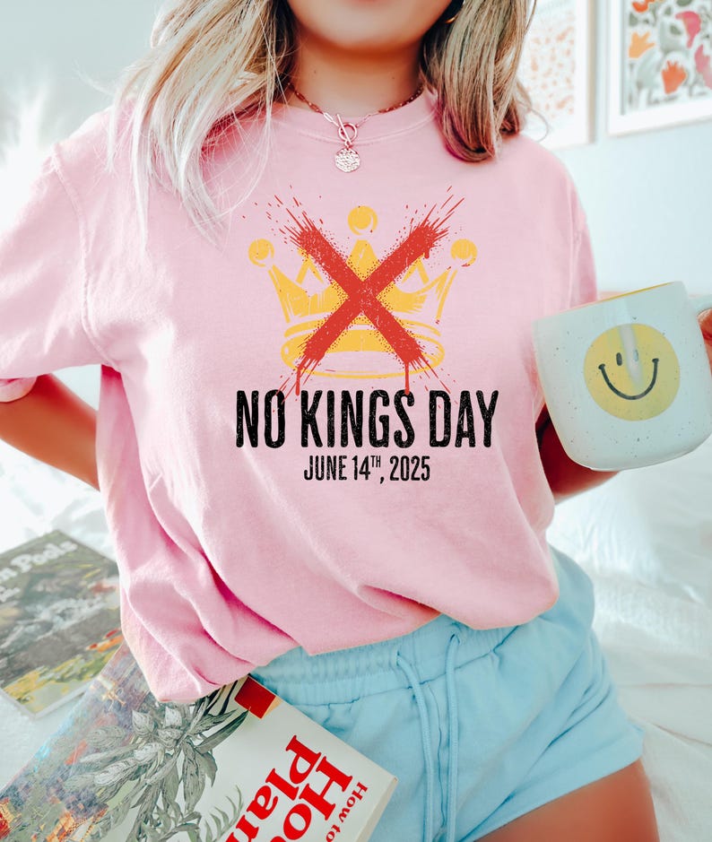 May include: Pink t-shirt with the text "NO KINGS DAY" and the date "JUNE 14TH, 2025". The design includes a yellow crown crossed out with a red "X". A white mug with a yellow smiley face is held in the hand.