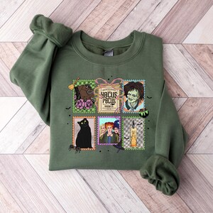 Puede incluir: Una sudadera verde oliva con un gr&aacute;fico colorido de sellos postales con im&aacute;genes y texto de la pel&iacute;cula "Hocus Pocus". Los sellos incluyen un gato negro, un caballo y las hermanas Sanderson.