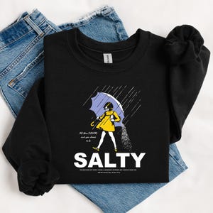May include: Black sweatshirt with a graphic of a person under a blue umbrella, with the word "SALTY" in white. The person is wearing a yellow coat and the text "All the flavors and you choose to be" is above the word "SALTY".