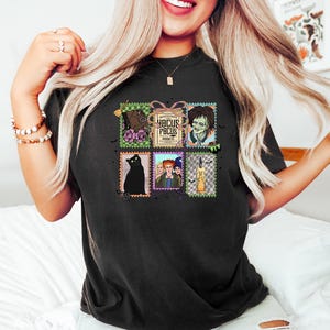 Puede incluir: Camiseta negra con un collage de im&aacute;genes estilo sello postal. Las im&aacute;genes incluyen un gato negro, un zombi, una escoba y el texto "Hocus Pocus". La camiseta tiene cuello redondo y mangas cortas.
