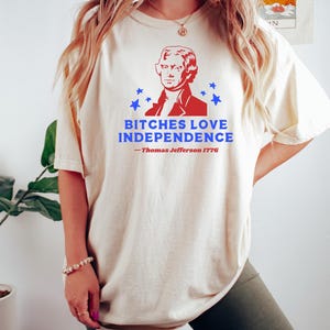 May include: Cream-colored t-shirt featuring a red and blue graphic of Thomas Jefferson and the text "BITCHES LOVE INDEPENDENCE - Thomas Jefferson 1776". The shirt has short sleeves and a relaxed fit.