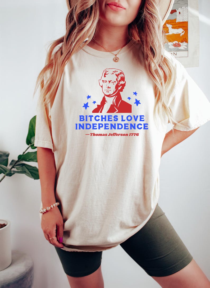 May include: Cream-colored t-shirt featuring a red and blue graphic of Thomas Jefferson and the text "BITCHES LOVE INDEPENDENCE - Thomas Jefferson 1776". The shirt has short sleeves and a relaxed fit.