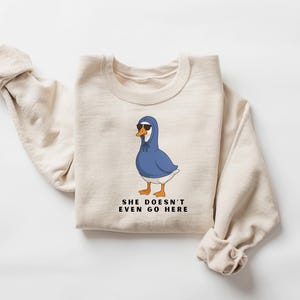 May include: A cream-coloured sweatshirt with a cartoon goose wearing a blue hoodie and sunglasses. The goose has orange feet and a white chest. The text below the goose reads, "SHE DOESN'T EVEN GO HERE."