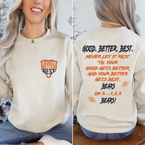 May include: Cream-coloured sweatshirt. The front features the text "GOOD BETTER REST" in a shield design. The back displays the text "GOOD. BETTER. BEST. NEVER LET IT REST... BEARS ON 3...1,2,3 BEARS!" in navy and orange.
