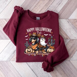 May include: A maroon sweatshirt featuring the phrase "HAPPY HALLOWEENIE Y'all" above a cartoon image of a dachshund and a ghost dressed as cowboys. The dachshund is wearing a cowboy hat and bandana, with pumpkins and cacti.