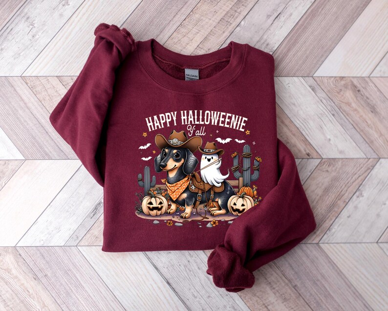 May include: A maroon sweatshirt featuring the phrase "HAPPY HALLOWEENIE Y'all" above a cartoon image of a dachshund and a ghost dressed as cowboys. The dachshund is wearing a cowboy hat and bandana, with pumpkins and cacti.