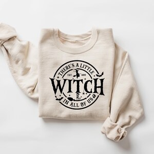 Op de afbeelding: Beige sweatshirt met een ronde hals en lange mouwen. De voorkant heeft een zwart cirkelvormig ontwerp met de tekst "THERE'S A LITTLE WITCH IN ALL OF US" en heksenillustraties. De tekst bevat "EST. 1692".