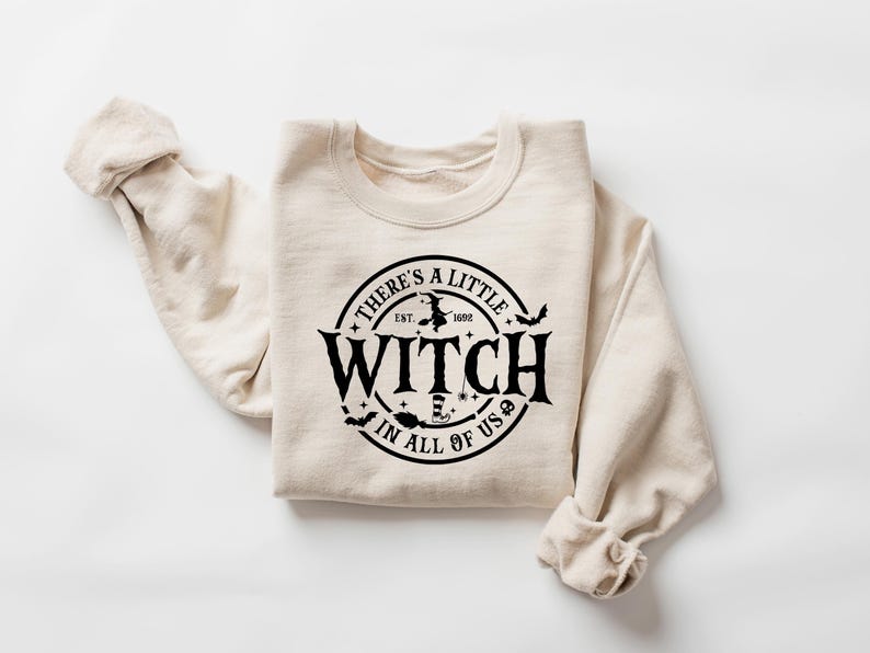 Op de afbeelding: Beige sweatshirt met een ronde hals en lange mouwen. De voorkant heeft een zwart cirkelvormig ontwerp met de tekst "THERE'S A LITTLE WITCH IN ALL OF US" en heksenillustraties. De tekst bevat "EST. 1692".