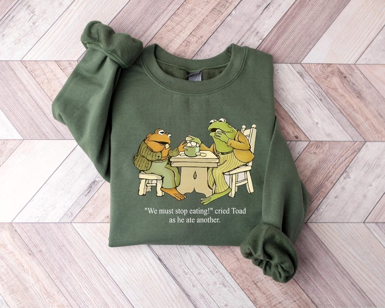 Puede incluir: Sudadera verde con una ilustraci&oacute;n de dibujos animados de dos ranas en una mesa. Las ranas visten ropa y est&aacute;n comiendo. El texto de la sudadera dice: "&iexcl;Debemos dejar de comer!" grit&oacute; Sapo mientras com&iacute;a otro.
