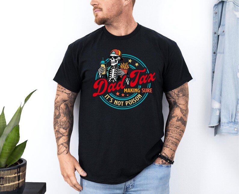 Pode incluir: Camiseta preta com um design gr&aacute;fico circular com um esqueleto usando um bon&eacute;, segurando uma bebida e batatas fritas. O texto diz "Dad Tax making sure it's not poison" em vermelho e branco. O design inclui estrelas e uma borda azul.