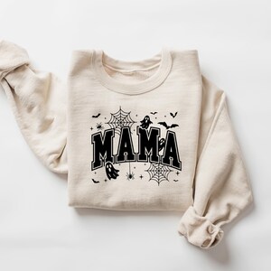 Op de afbeelding: Een beige sweatshirt met het woord "MAMA" in vette, zwarte letters. Het ontwerp bevat zwarte spinnenwebben, spinnen, vleermuizen en geesten, wat een Halloween-thema cre&euml;ert. De mouwen zijn opgerold.