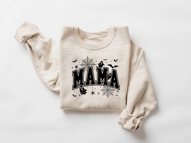 Op de afbeelding: Een beige sweatshirt met het woord "MAMA" in vette, zwarte letters. Het ontwerp bevat zwarte spinnenwebben, spinnen, vleermuizen en geesten, wat een Halloween-thema cre&euml;ert. De mouwen zijn opgerold.