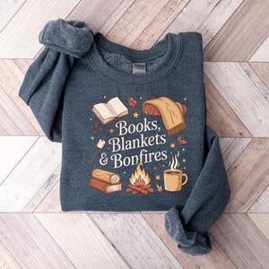 May include: A navy blue sweatshirt with the words "Books, Blankets & Bonfires" in a whimsical font. The graphic includes illustrations of a book, blanket, campfire, mug, and marshmallows. The sweatshirt is folded on a light wood-grain surface.