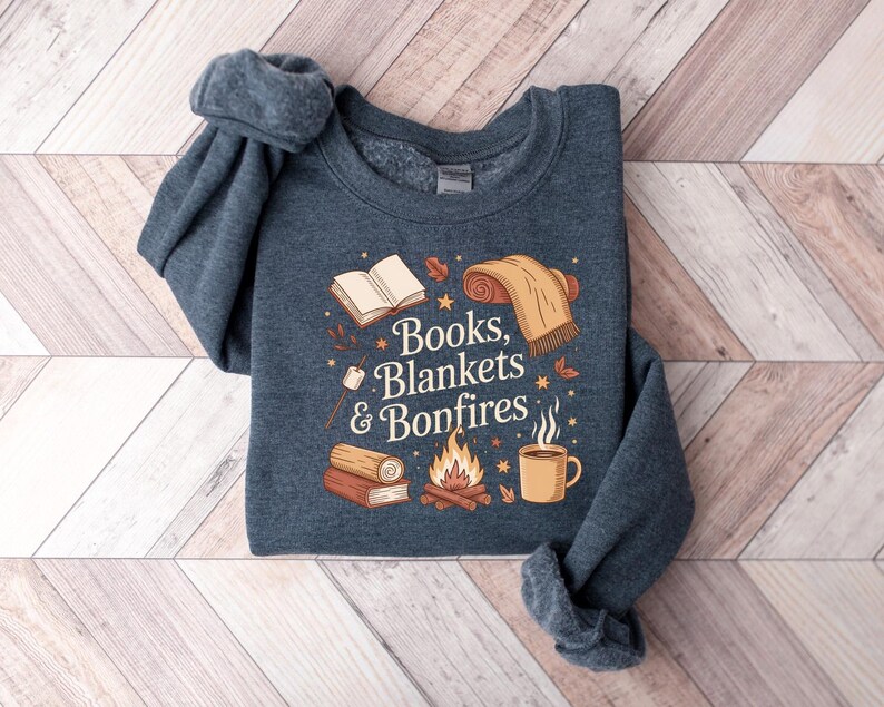 May include: A navy blue sweatshirt with the words "Books, Blankets & Bonfires" in a whimsical font. The graphic includes illustrations of a book, blanket, campfire, mug, and marshmallows. The sweatshirt is folded on a light wood-grain surface.