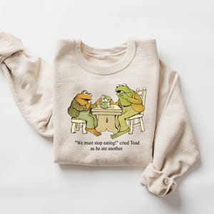 Puede incluir: Sudadera beige con una ilustraci&oacute;n de dibujos animados de dos ranas sentadas a una mesa. Las ranas visten ropa verde y marr&oacute;n. El texto en la sudadera dice: "&iexcl;Debemos dejar de comer!" grit&oacute; Sapo mientras com&iacute;a otro.