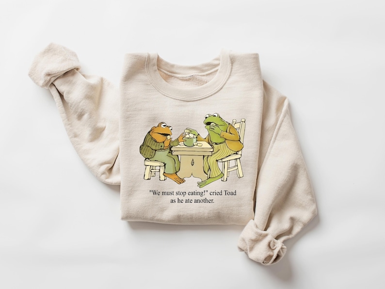 Puede incluir: Sudadera beige con una ilustraci&oacute;n de dibujos animados de dos ranas sentadas a una mesa. Las ranas visten ropa verde y marr&oacute;n. El texto en la sudadera dice: "&iexcl;Debemos dejar de comer!" grit&oacute; Sapo mientras com&iacute;a otro.