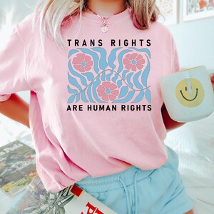 May include: Pink t-shirt featuring a graphic design of blue and pink flowers and leaves. The text "Trans Rights Are Human Rights" is printed on the shirt.