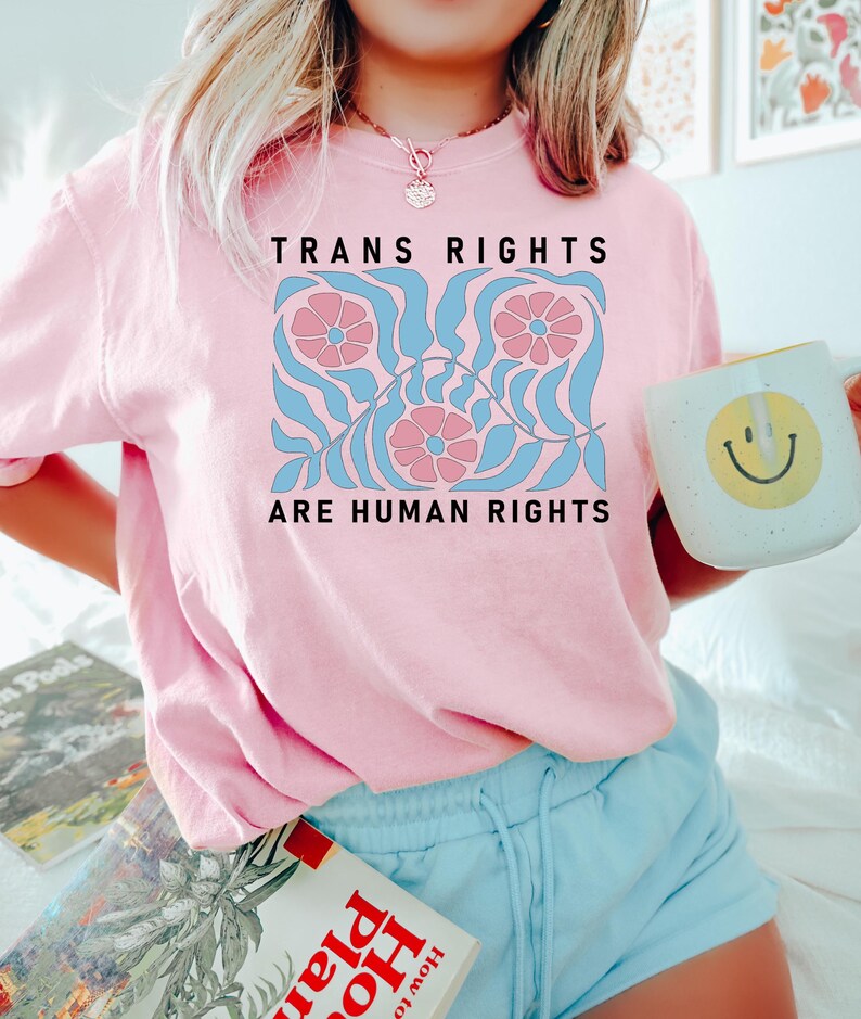 May include: Pink t-shirt featuring a graphic design of blue and pink flowers and leaves. The text "Trans Rights Are Human Rights" is printed on the shirt.