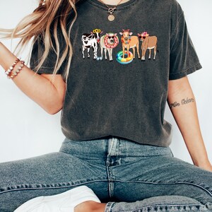 Pu&ograve; includere: T-shirt grigio scuro con una grafica di cinque mucche dei cartoni animati con occhiali da sole e accessori estivi. Le mucche sono di vari colori, tra cui bianco e nero, marrone e beige. La maglietta &egrave; abbinata a jeans blu e scarpe da ginnastica bianche.