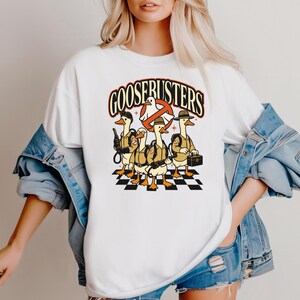 May include: White t-shirt featuring a cartoon graphic of five geese dressed as Ghostbusters, with the text "GOOSEBUSTERS" above them. The geese stand on a black and white checkered surface.