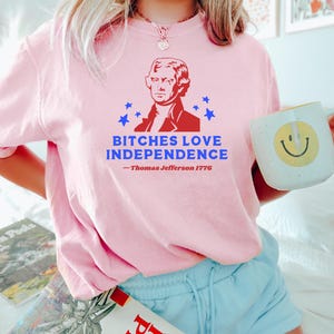 May include: A light pink t-shirt with the text "BITCHES LOVE INDEPENDENCE" and a portrait of Thomas Jefferson. The shirt also includes the text "- Thomas Jefferson 1776". The person is holding a white mug with a yellow smiley face.