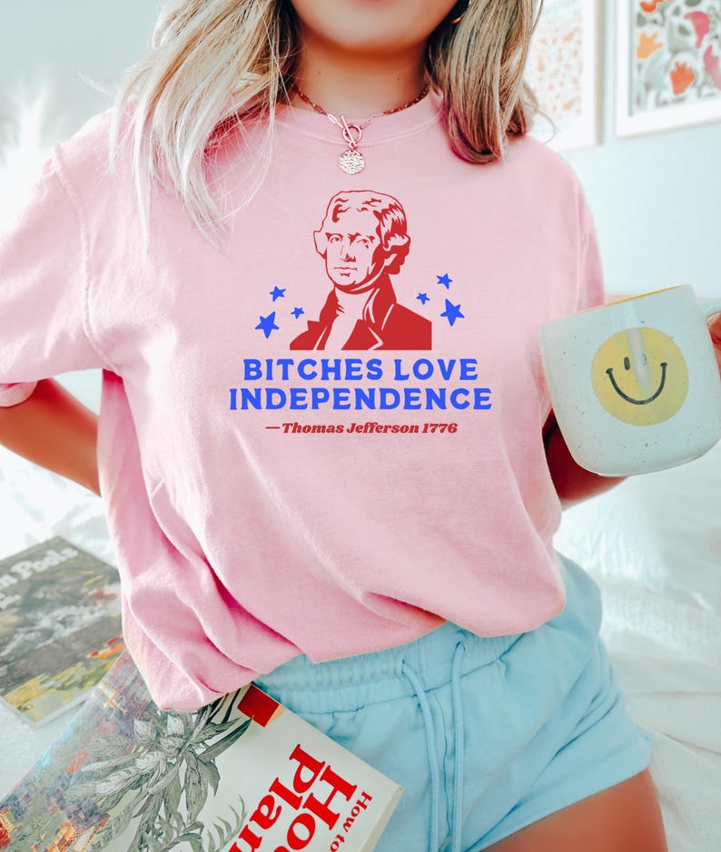 May include: A light pink t-shirt with the text "BITCHES LOVE INDEPENDENCE" and a portrait of Thomas Jefferson. The shirt also includes the text "- Thomas Jefferson 1776". The person is holding a white mug with a yellow smiley face.