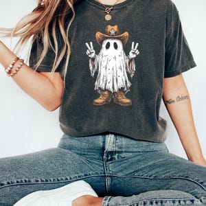 May include: Dark gray t-shirt featuring a cartoon ghost wearing a cowboy hat and boots, giving peace signs. The ghost has a white sheet-like body with black eyes. The shirt is paired with blue jeans and white shoes.