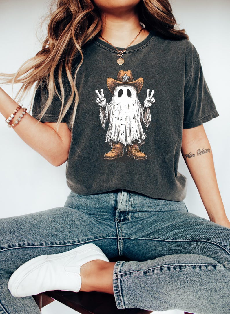 May include: Dark gray t-shirt featuring a cartoon ghost wearing a cowboy hat and boots, giving peace signs. The ghost has a white sheet-like body with black eyes. The shirt is paired with blue jeans and white shoes.