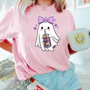 K&ouml;nnte beinhalten: Hellrosa T-Shirt mit einem Cartoon-Geist-Design. Der Geist hat eine lila Schleife und h&auml;lt eine Getr&auml;nkedose mit dem Text "Alan". Lila Sterne umgeben den Geist. Eine wei&szlig;e Tasse mit einem gelben Smiley ist ebenfalls sichtbar.