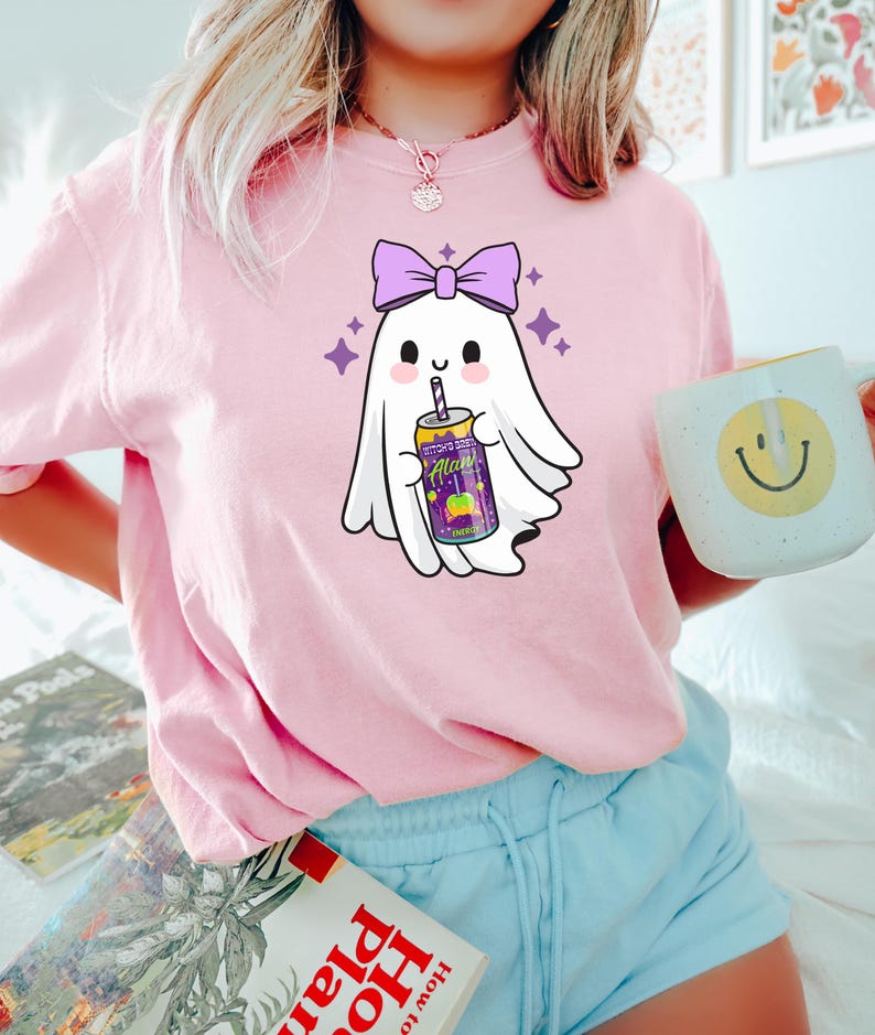 K&ouml;nnte beinhalten: Hellrosa T-Shirt mit einem Cartoon-Geist-Design. Der Geist hat eine lila Schleife und h&auml;lt eine Getr&auml;nkedose mit dem Text "Alan". Lila Sterne umgeben den Geist. Eine wei&szlig;e Tasse mit einem gelben Smiley ist ebenfalls sichtbar.