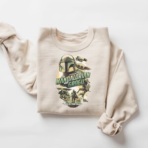 May include: A beige sweatshirt featuring a graphic from "The Mandalorian" with the text "The Mandalorian and Grogu". The design includes images of the Mandalorian helmet, Grogu, and other characters. The sweatshirt is folded.